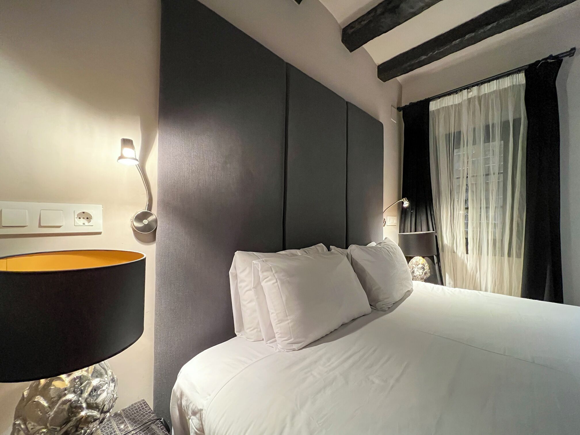 A hotel room with a large bed, a bedside lamp, a wall socket, and a window with sheer curtains.
