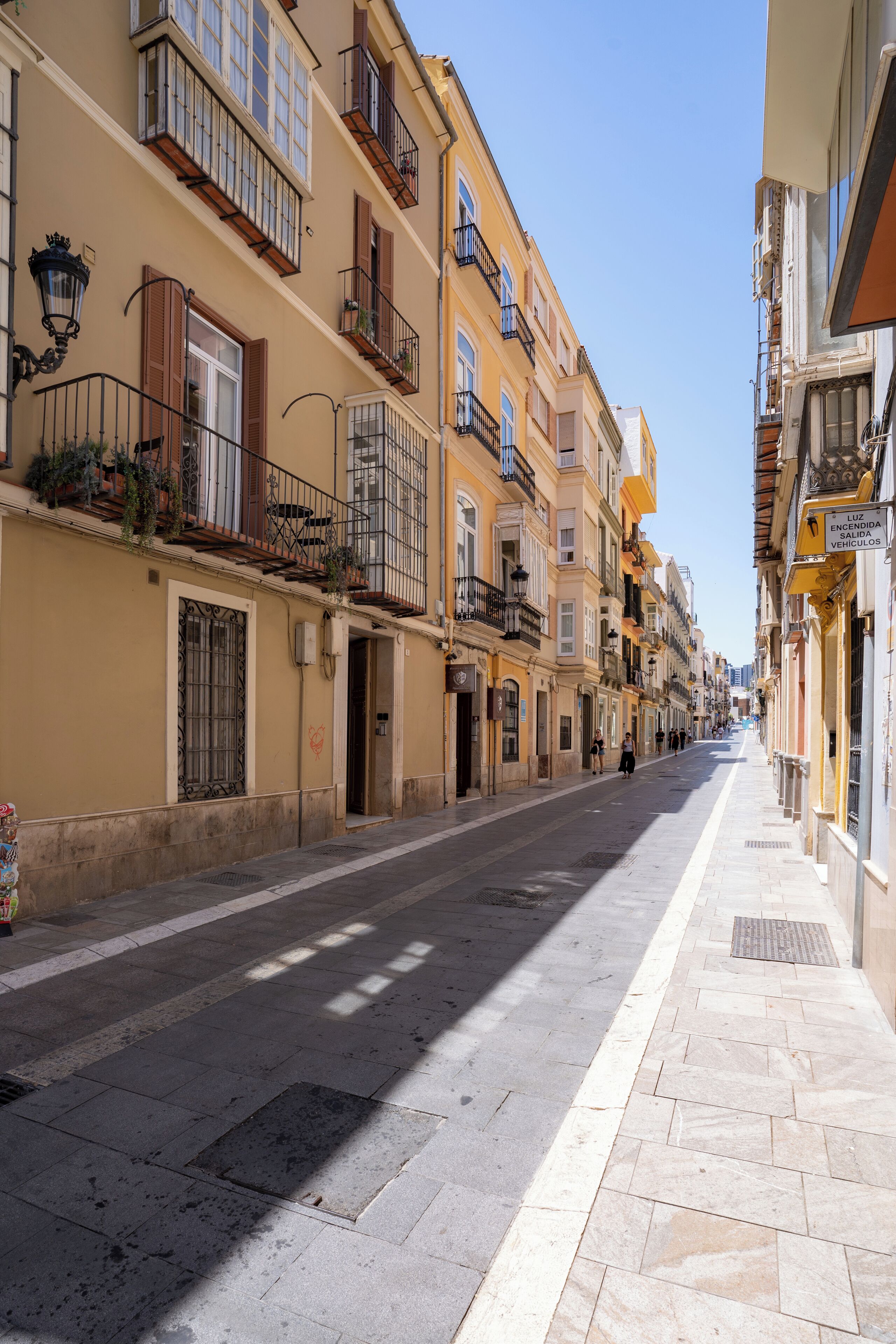 A street lined with multi-story buildings, featuring balconies with railings and potted plants.
