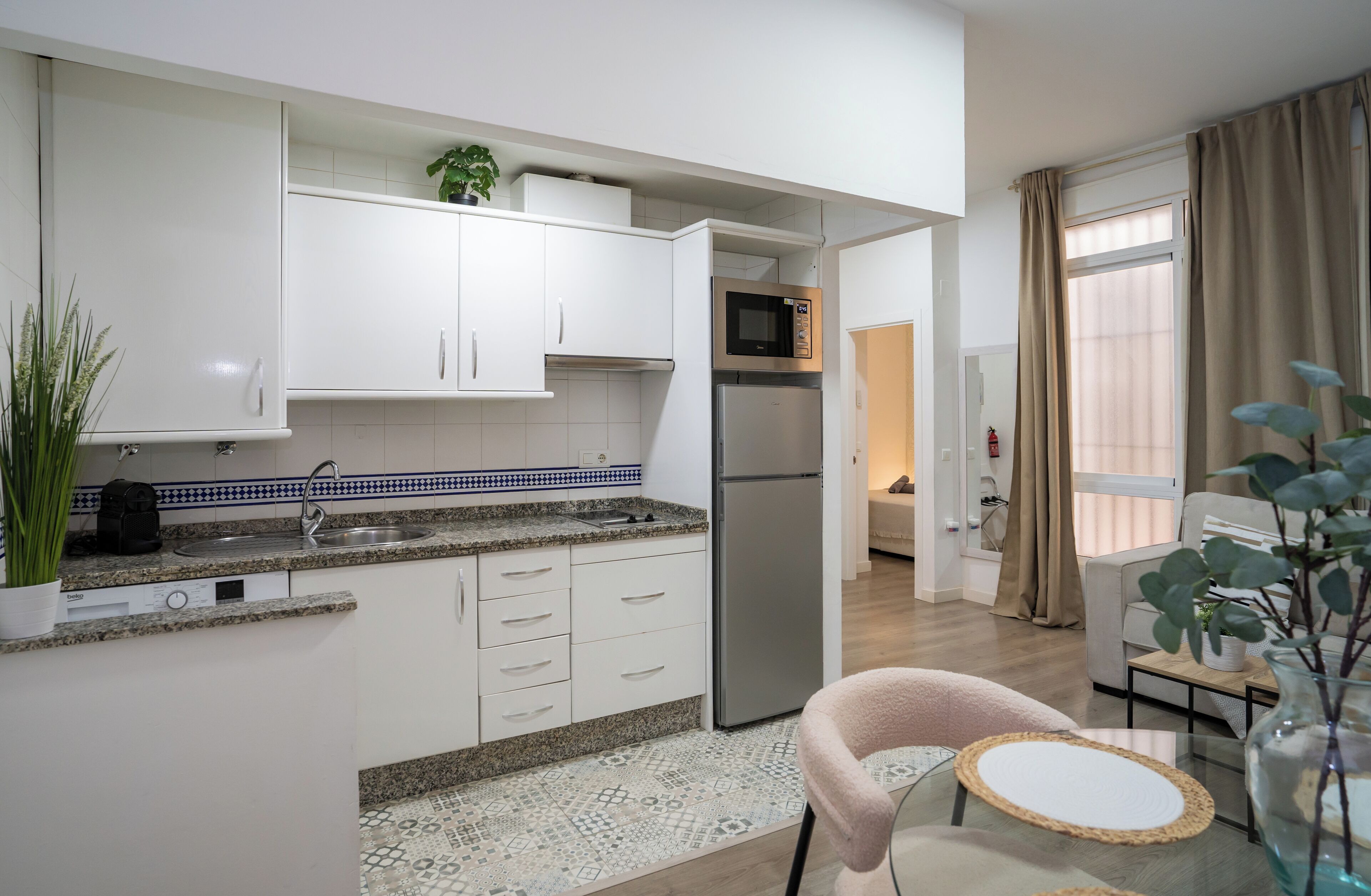 Una cocina moderna con armarios blancos, un refrigerador de acero inoxidable, un microondas y un lavamanos. Hay una pequeña zona de comedor con una silla y una mesa.