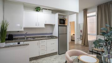 Una cocina moderna con armarios blancos, un refrigerador de acero inoxidable, un microondas y un lavamanos. Hay una pequeña zona de comedor con una silla y una mesa.