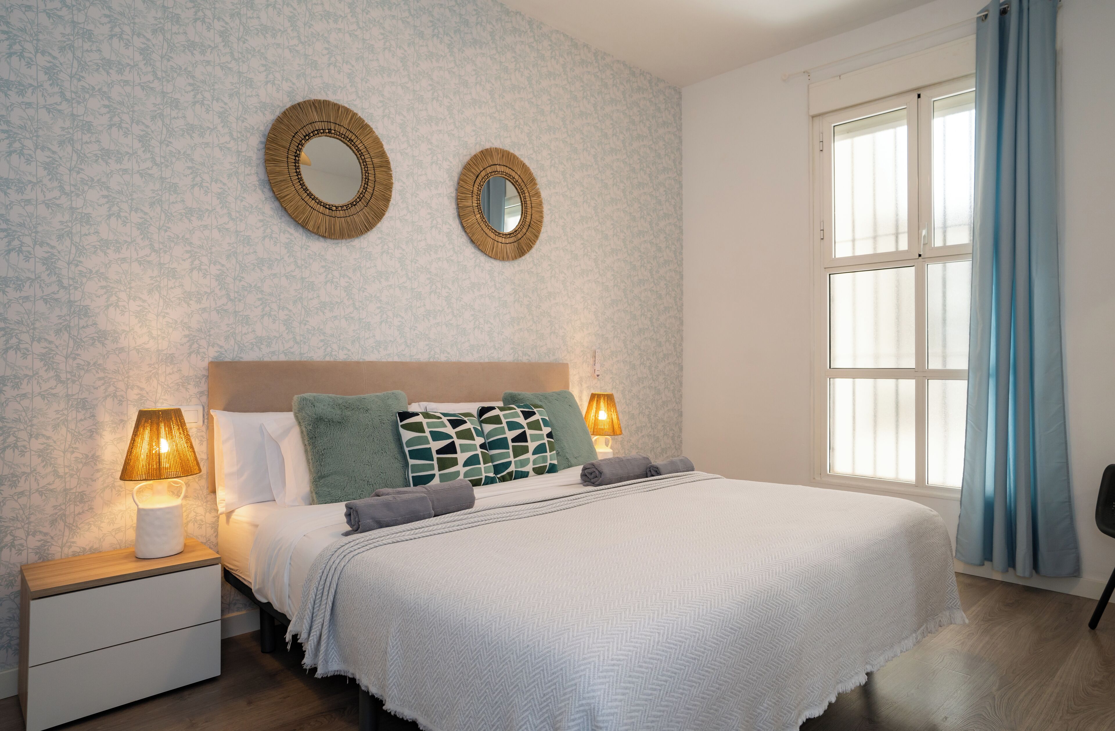 A bedroom with a bed, two bedside lamps, a nightstand, and a window with curtains.