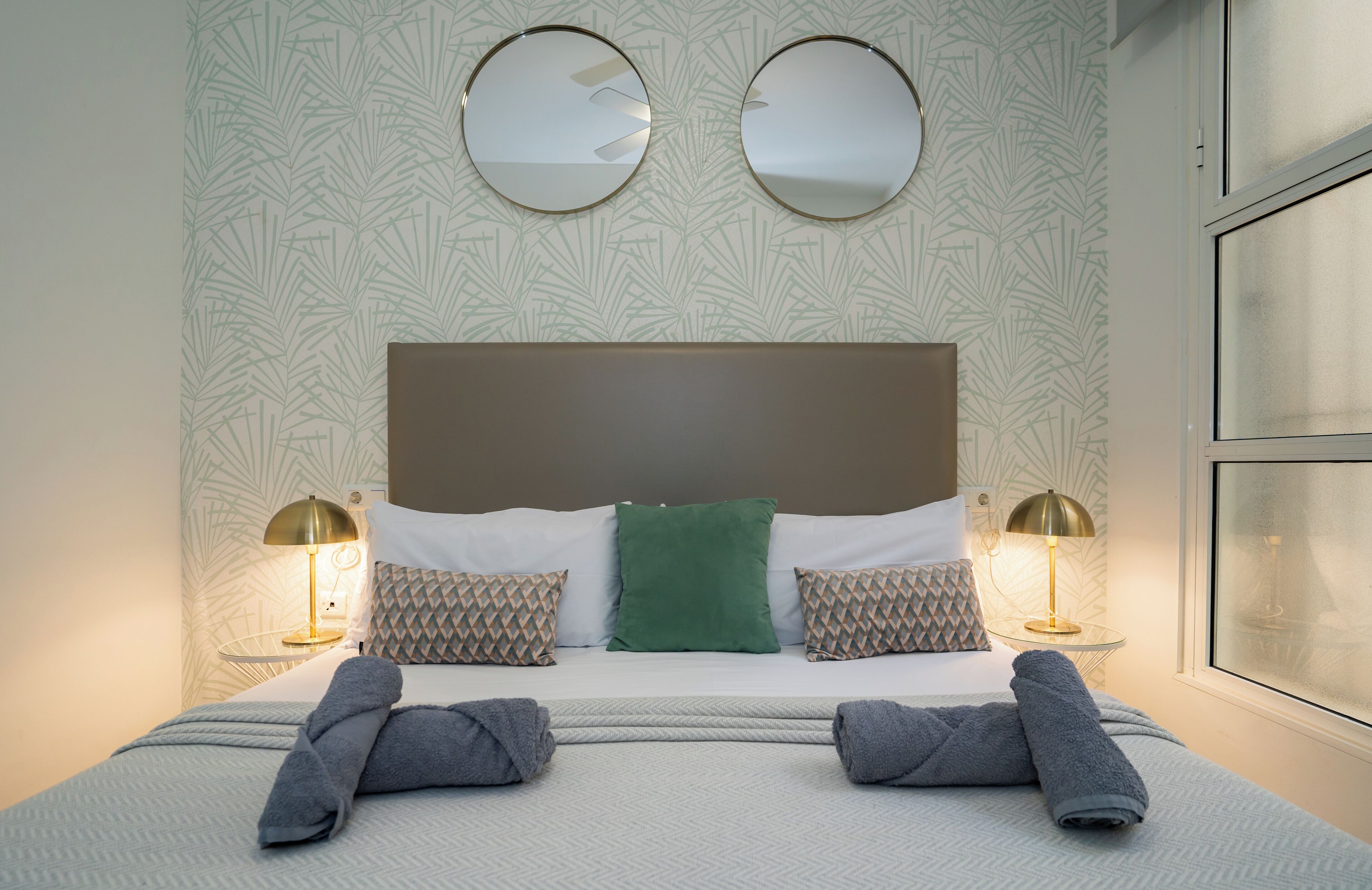 A neatly made bed with two pillows, two round mirrors above it, and two bedside lamps.