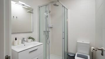 Une salle de bain moderne avec une douche à parois de verre, un lavabo blanc agrémenté d’une plante et des toilettes sur lesquelles est posée une serviette.