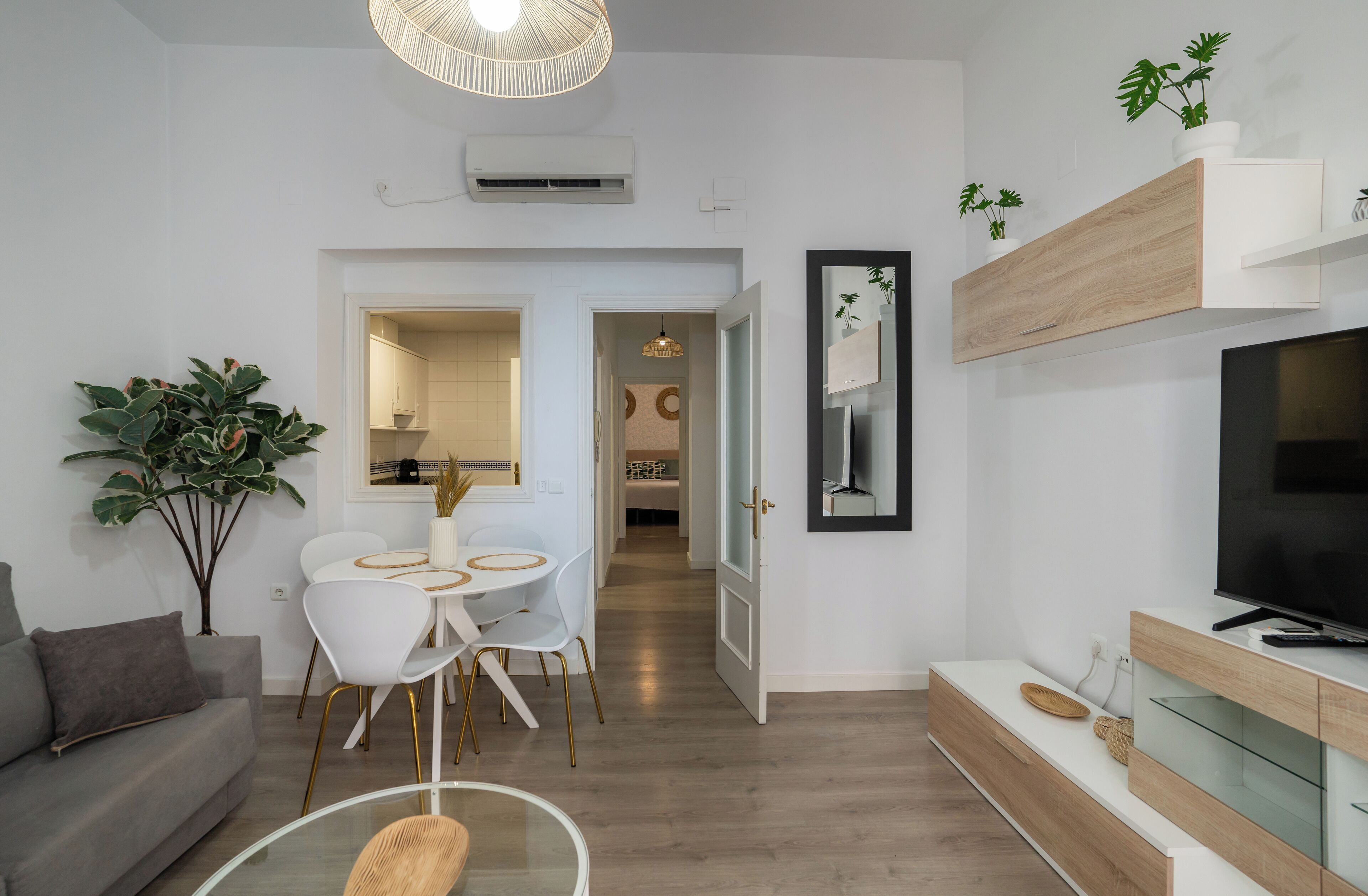 Una sala de estar moderna con un sofá gris, un comedor con una mesa y sillas blancas, un televisor sobre un mueble de madera y una planta grande en maceta.