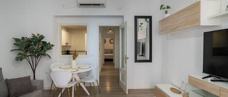 Una sala de estar moderna con un sofá gris, un comedor con una mesa y sillas blancas, un televisor sobre un mueble de madera y una planta grande en maceta.