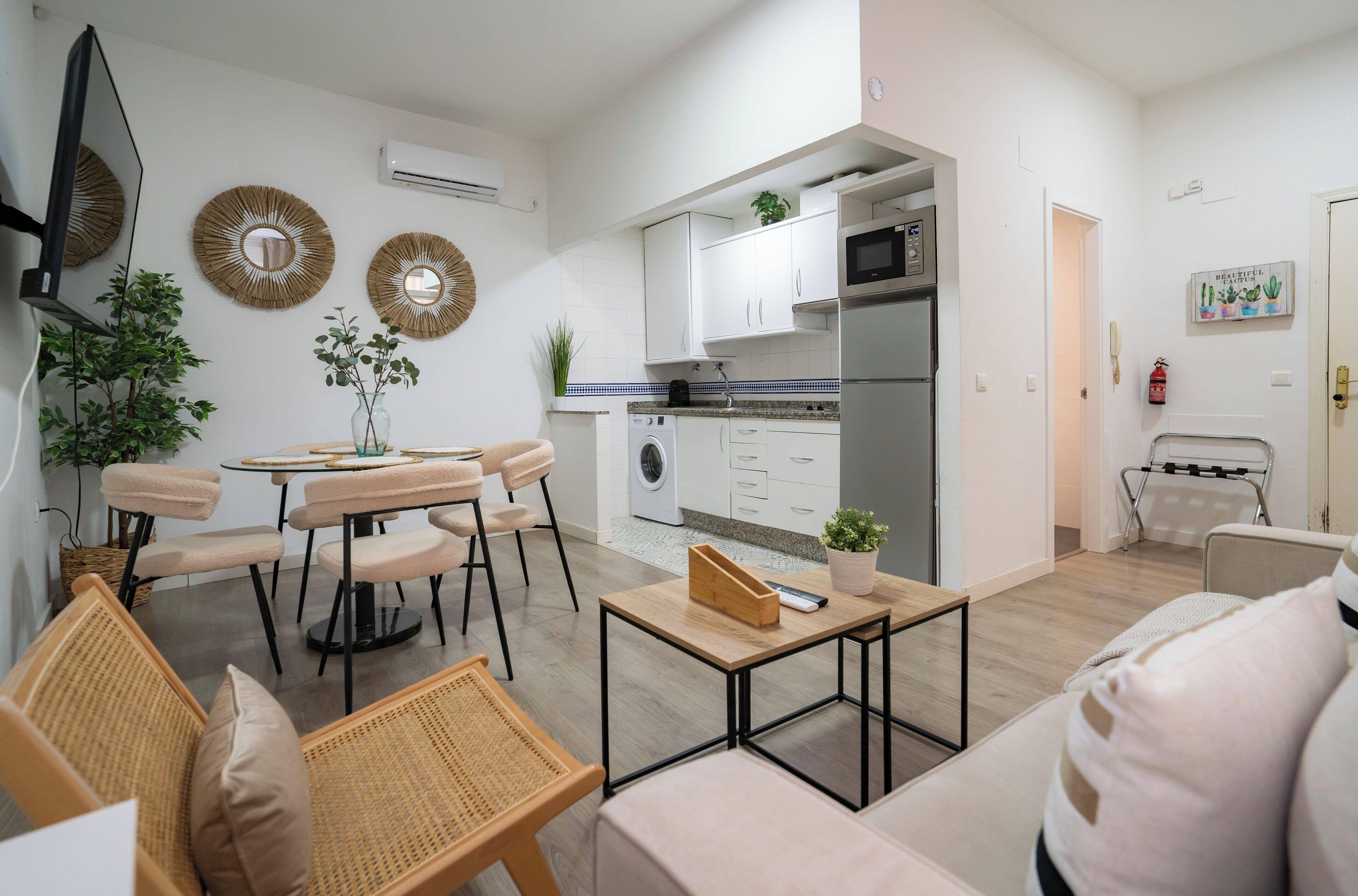 Una cocina moderna con armarios blancos, microondas y lavadora. Hay un comedor con mesa y sillas, y una sala de estar con sofá y televisor.