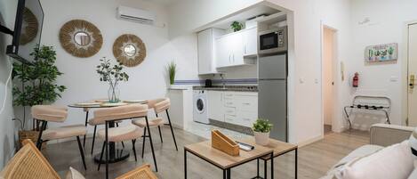 Una cocina moderna con armarios blancos, microondas y lavadora. Hay un comedor con mesa y sillas, y una sala de estar con sofá y televisor.