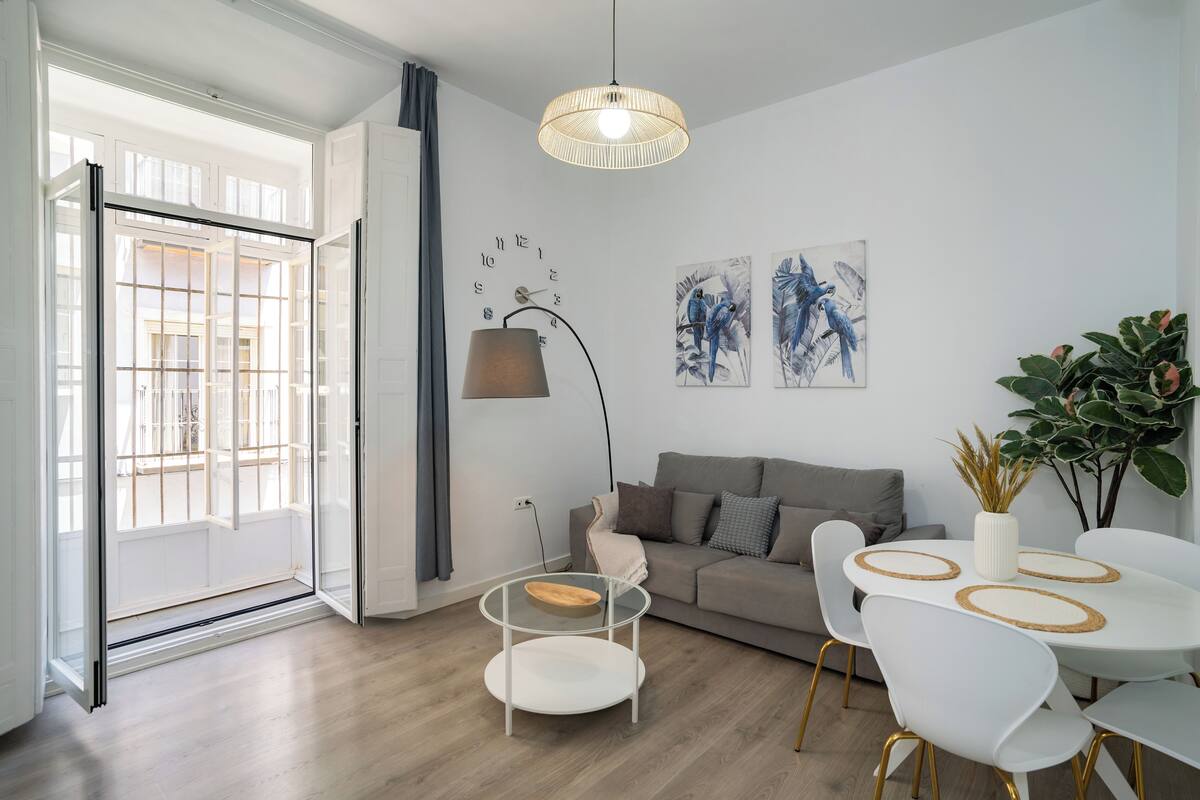 Ein modernes Wohnzimmer mit Glastür zum Balkon, einem runden Esstisch mit weißen Stühlen, einer Couch und einer Stehlampe.