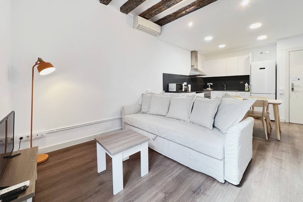 Un salon moderne avec un canapé blanc, une petite table en bois et un lampadaire. On y trouve également un coin cuisine avec des meubles blancs et un four à micro-ondes.