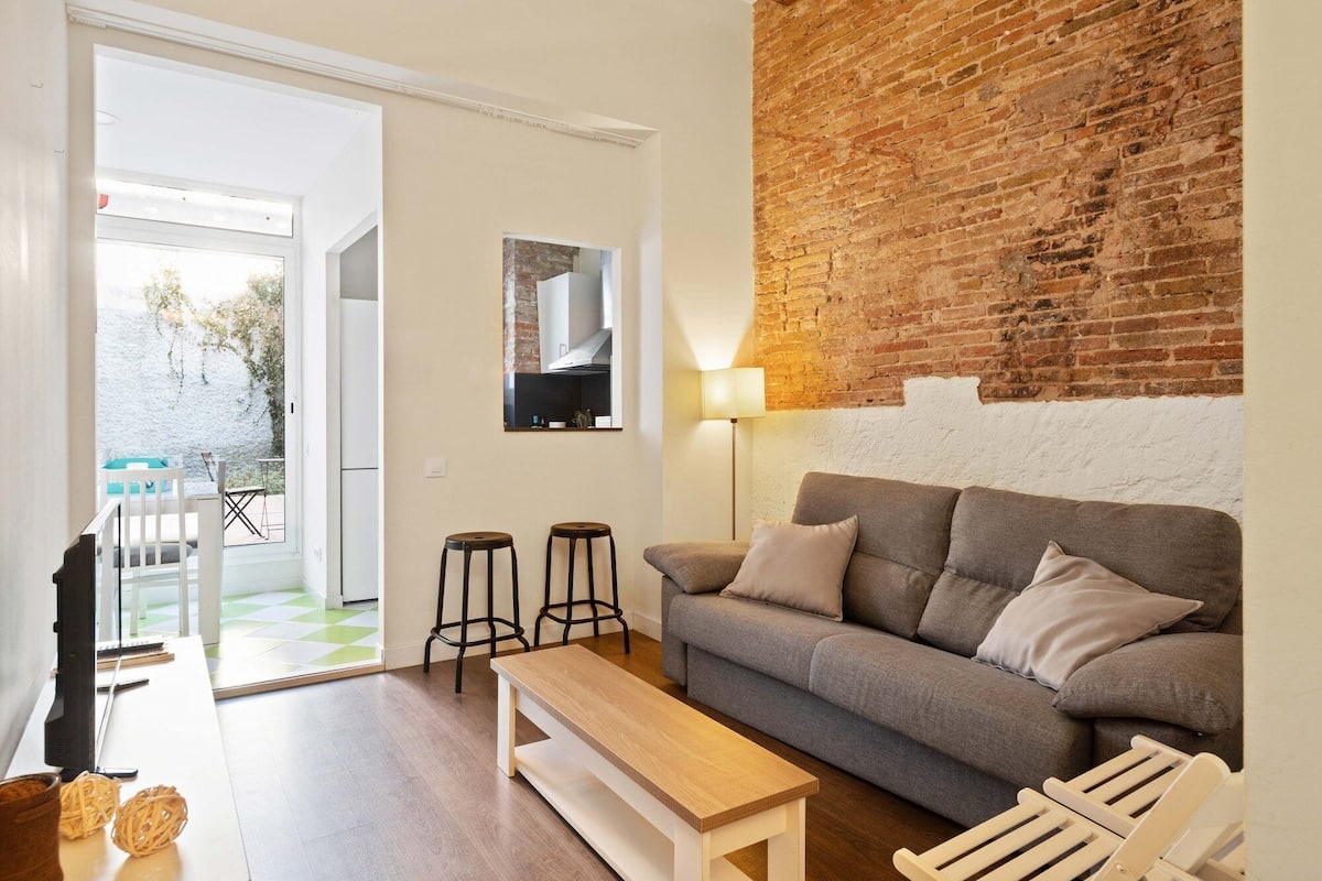 Un salon avec un canapé gris, deux tabourets de bar noirs, une table basse en bois, un téléviseur et une lampe. Il y a un mur en briques et une porte blanche qui donne sur l’extérieur.