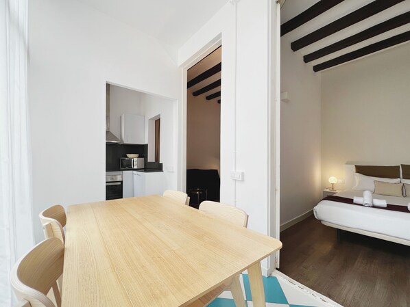 A compact hotel room with a wooden dining table, a small kitchenette, a bed with white bedding, and a nightstand with a lamp.