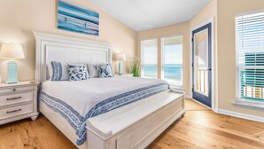 A bedroom with a large bed, a nightstand, a lamp, and a window with blinds. - Ocean Breeze Beachfront SGI: Relaxing Escape for Families & Couples with Pool (St. George Island)