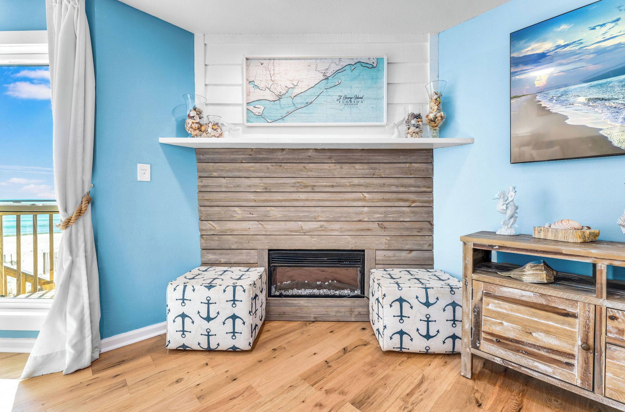Ein Zimmer mit blauer Wand, Holzboden und Kamin. Zwei passende Hocker mit Anker-Motiven, eine Karte über dem Kamin und ein Bild von einer Strandansicht.