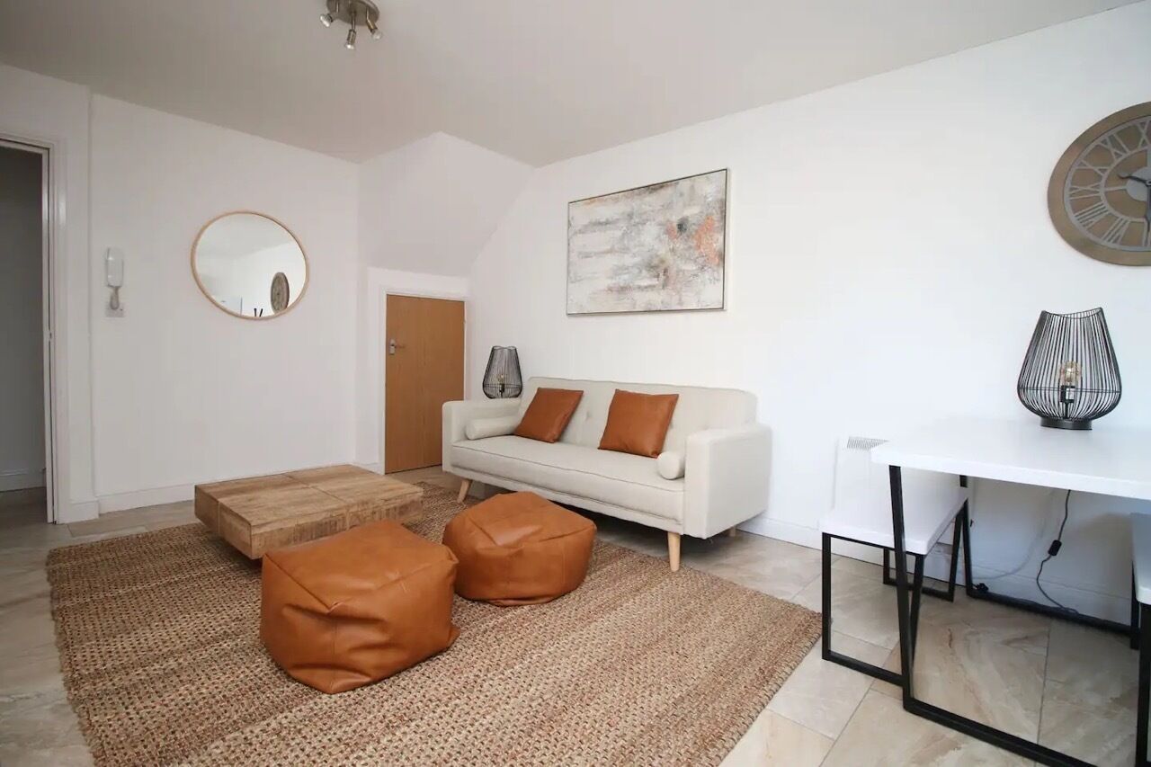 Una sala moderna con un sofá blanco, dos otomanas marrones, un espejo redondo, un cuadro y una mesa con un ventilador.