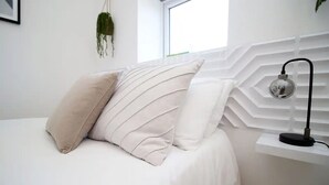 Un lit bien fait, recouvert d’une couvre-lit blanche et agrémenté de deux oreillers, une plante suspendue et un luminaire fixé au mur.