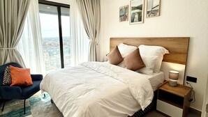 A modern bedroom with a large bed, a nightstand with a lamp, a view of the city through the window, and a colorful rug. - 1-Bedroom Pool View Unit @Nova (Accra)