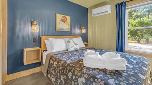 A bedroom with a bed, a nightstand, a wall-mounted lamp, a painting, and an air conditioning unit.