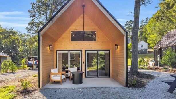 A wooden cabin with a pitched roof, large sliding glass doors, and a patio area with outdoor furniture. - Hot Tub! Riverfront Cabin Retreat - 6 Min to GLV (Easley)