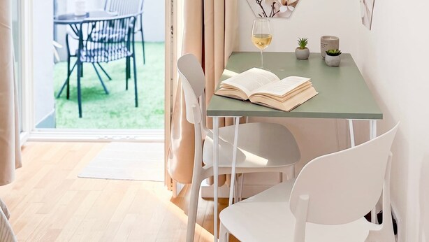 A bright room with a glass table, white chairs, a book, a glass of wine, and a view of an outdoor area with a table and chairs. - TurnKey | National Museum Studio with Patio I (Praha 1)