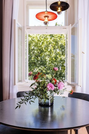 Une table ronde ornée d’un vase de fleurs au centre, une fenêtre donnant sur un paysage verdoyant et un luminaire suspendu au-dessus.
