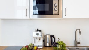 A modern kitchen with white cabinets, a stainless steel microwave, a coffee maker, a kettle, a sink, and fresh fruits on the counter. - TurnKey | National Museum Studio I-3 (Praha 1)