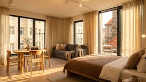 A modern hotel room with a large bed, a sofa, a dining area with a table and chairs, and large windows offering a city view. - TurnKey | Henry's Tower Studio with Terrace III-3 (Hlavní město Praha)