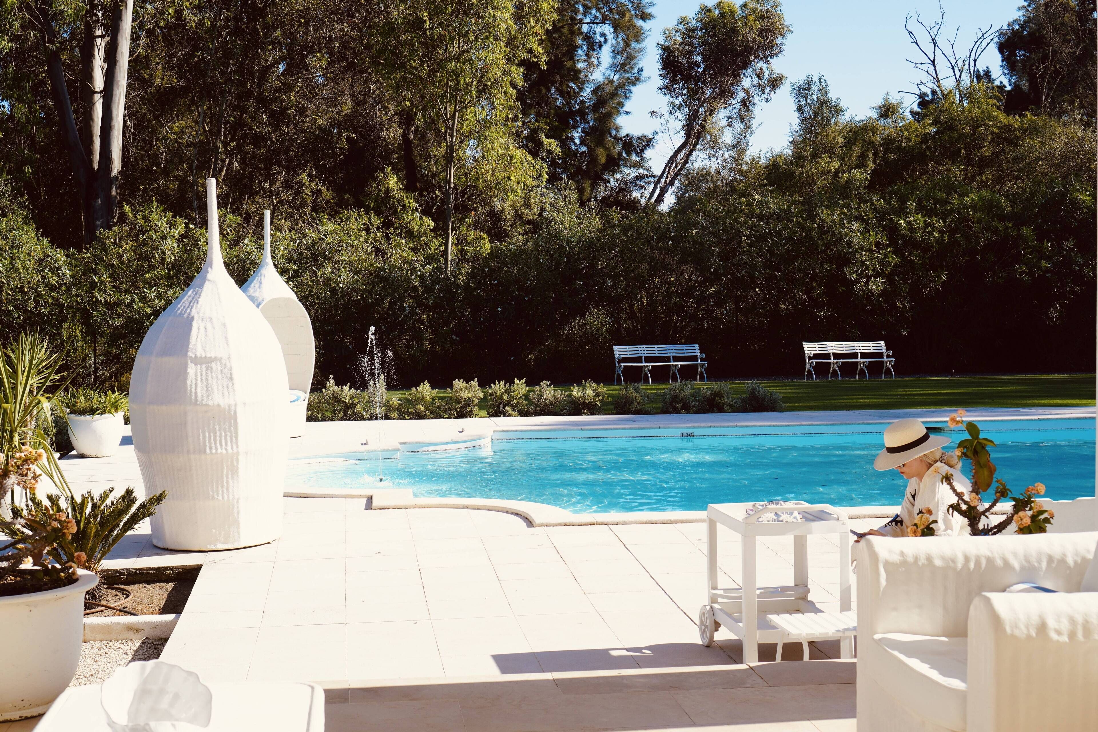 A poolside area with white outdoor furniture, a large white vase, and a bench in the background.