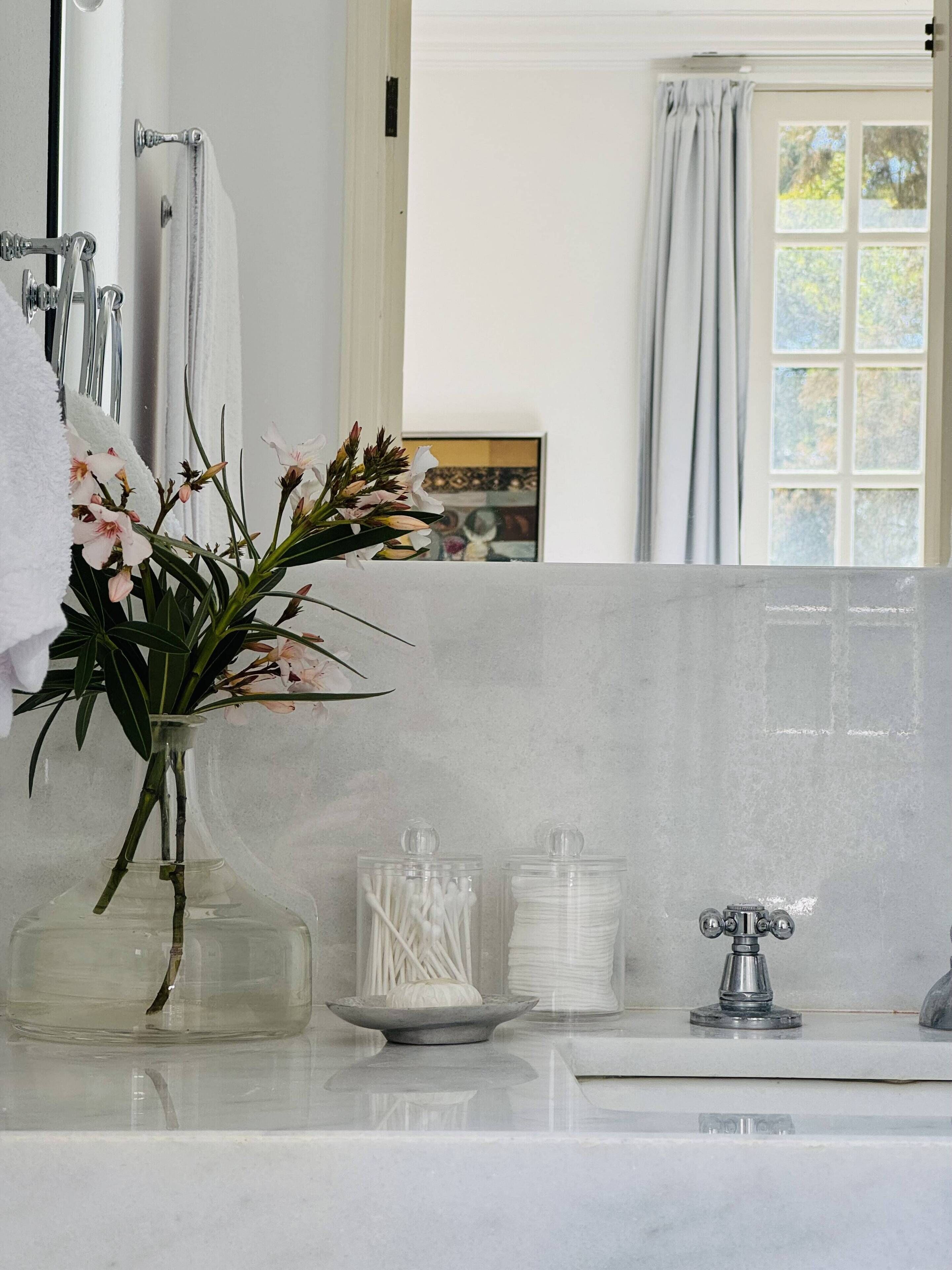 Ein Badezimmer mit einer weißen Marmorplatte, einer Glasvase mit Blumen, Wattestäbchen und einem Metallarmaturen.