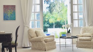 Una habitación con dos sillones color beige, una pequeña mesa redonda, una silla de madera oscura, un cuadro abstracto colorido y vistas de vegetación al exterior.