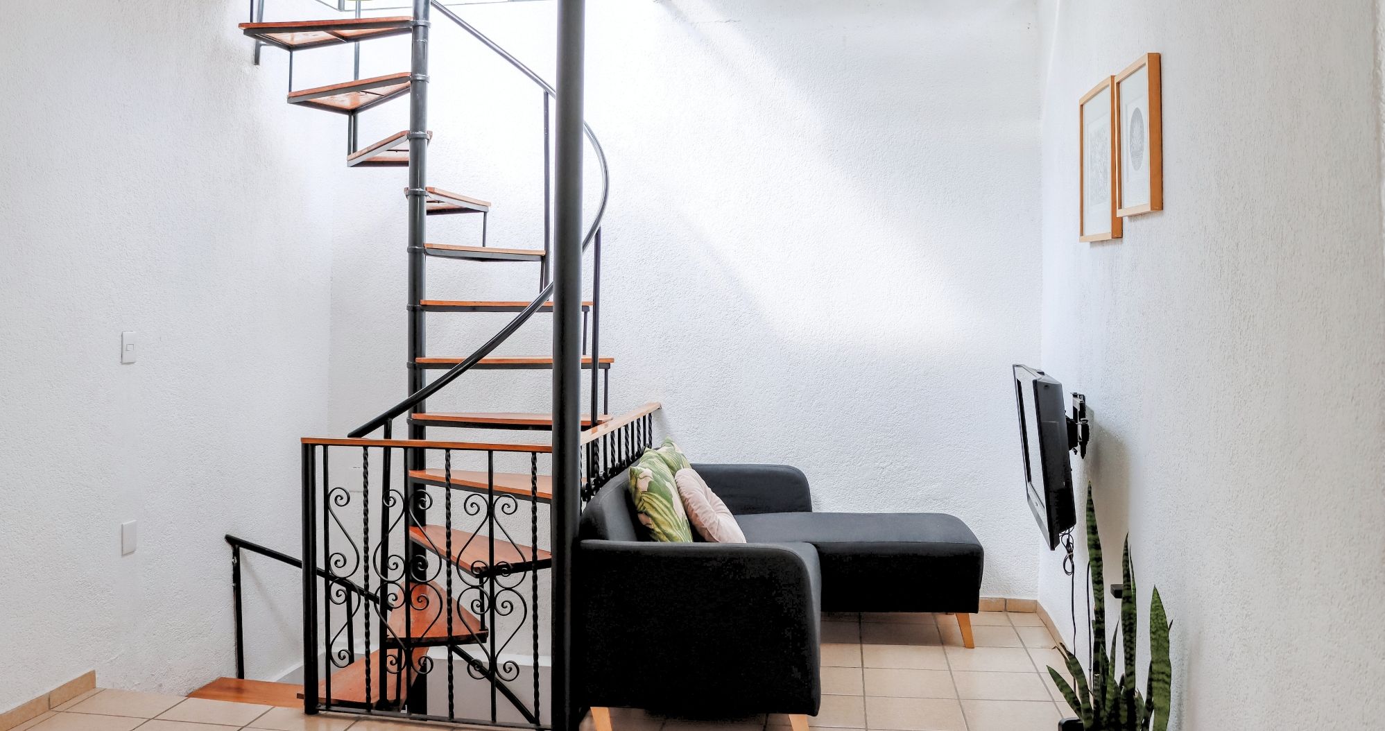 A compact living space with a spiral staircase, a black sofa, a TV, and a plant.