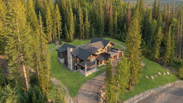 A modern two-story lodge surrounded by dense forest, with a clear view of the mountains in the background.