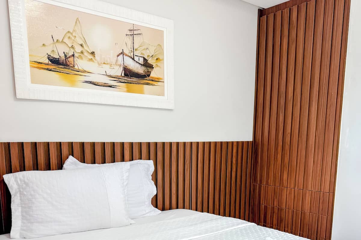 A neatly made bed with white linens and towels, a wooden headboard, and a painting of a boat on a river.