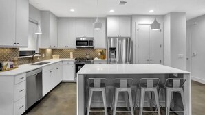 A modern kitchen with white cabinetry, stainless steel appliances, and a central island with bar stools. - Cityscape Retreat | Sleeps 14 | City Views + Grill (Austin)