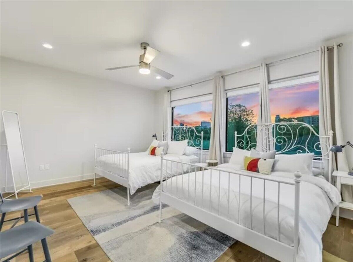 Un dormitorio moderno con dos camas de metal blanco, un escritorio y un ventanal con vista a la ciudad.