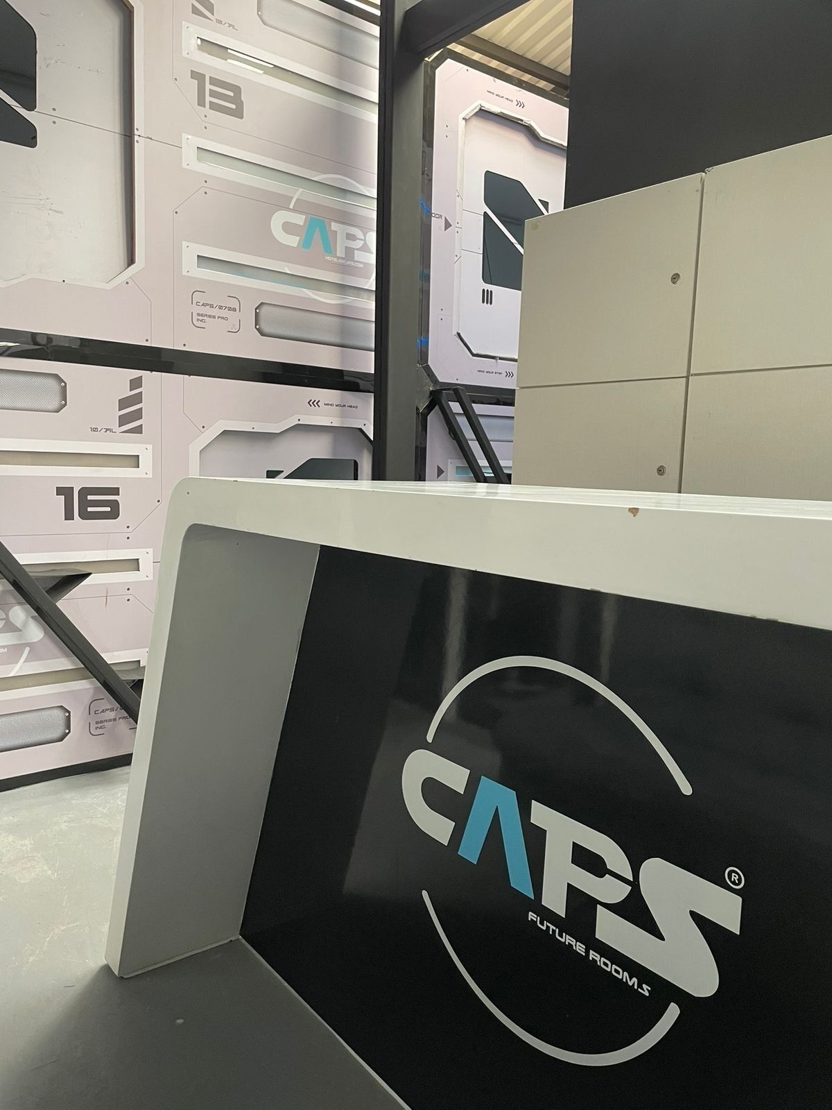 Une pièce futuriste ornée d’un logo bien visible indiquant « CAPS FUTURE ROOMS », d’une structure métallique et d’une échelle.