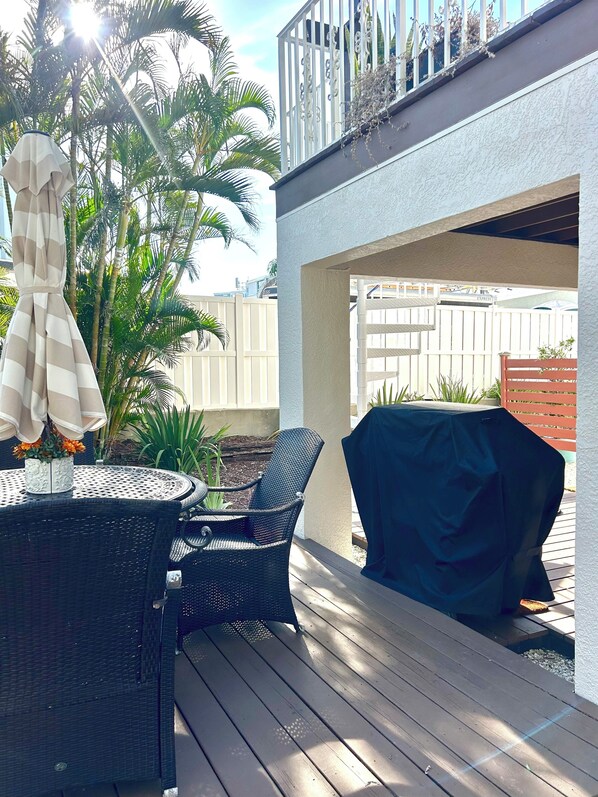Une terrasse avec une table ronde et des chaises, un barbecue abrité et, au fond, une clôture blanche.