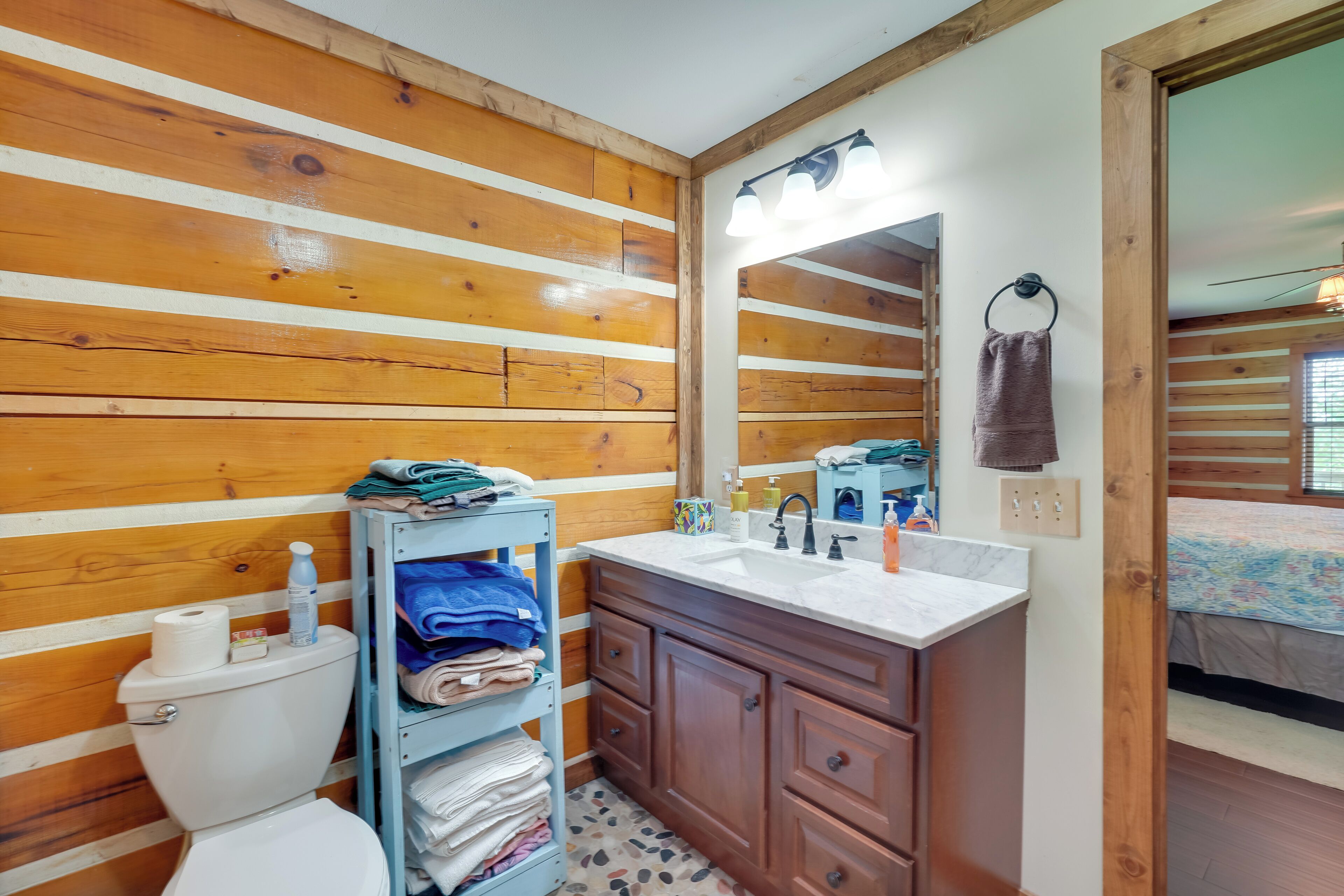A bathroom with wooden panel walls, a white toilet, a wooden vanity with a mirror, and a bed in the adjacent room.