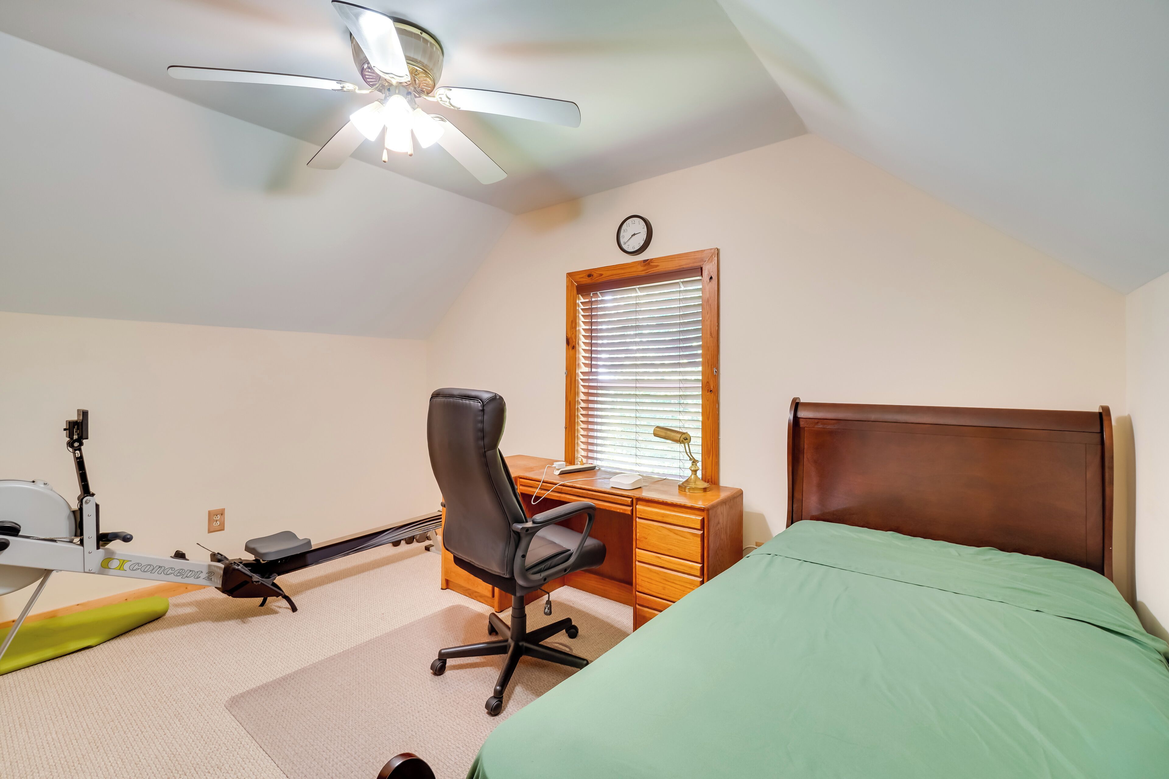 A bedroom with a bed, desk, chair, ceiling fan, and exercise equipment.