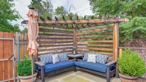 Een houten pergola met een blauwe, beklede bank en een tafel, omringd door potplanten en een patroonrijk vloerkleed.