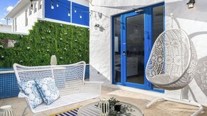 Un espace extérieur aménagé avec des sièges blancs, comprenant un canapé en osier blanc et un fauteuil suspendu, une table ronde agrémentée d’une plante, et une porte bleue donnant sur une pièce.