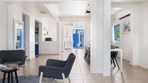 Un hall d’entrée spacieux au design moderne, comprenant un coin salon avec deux fauteuils et une petite table, un coin repas avec une table et des chaises, et une cuisine visible au fond.
