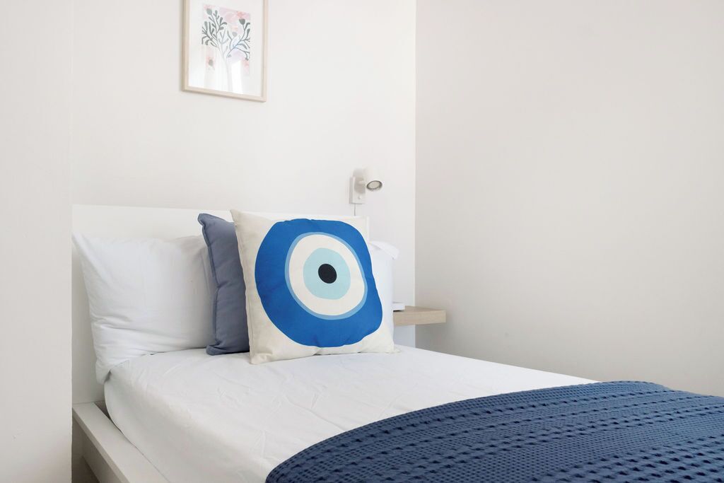 Een netjes opgemaakt bed met witte lakens en een blauw kussen in de vorm van een oog, een blauwe gebreide deken, en een ingelijst schilderij aan de muur.