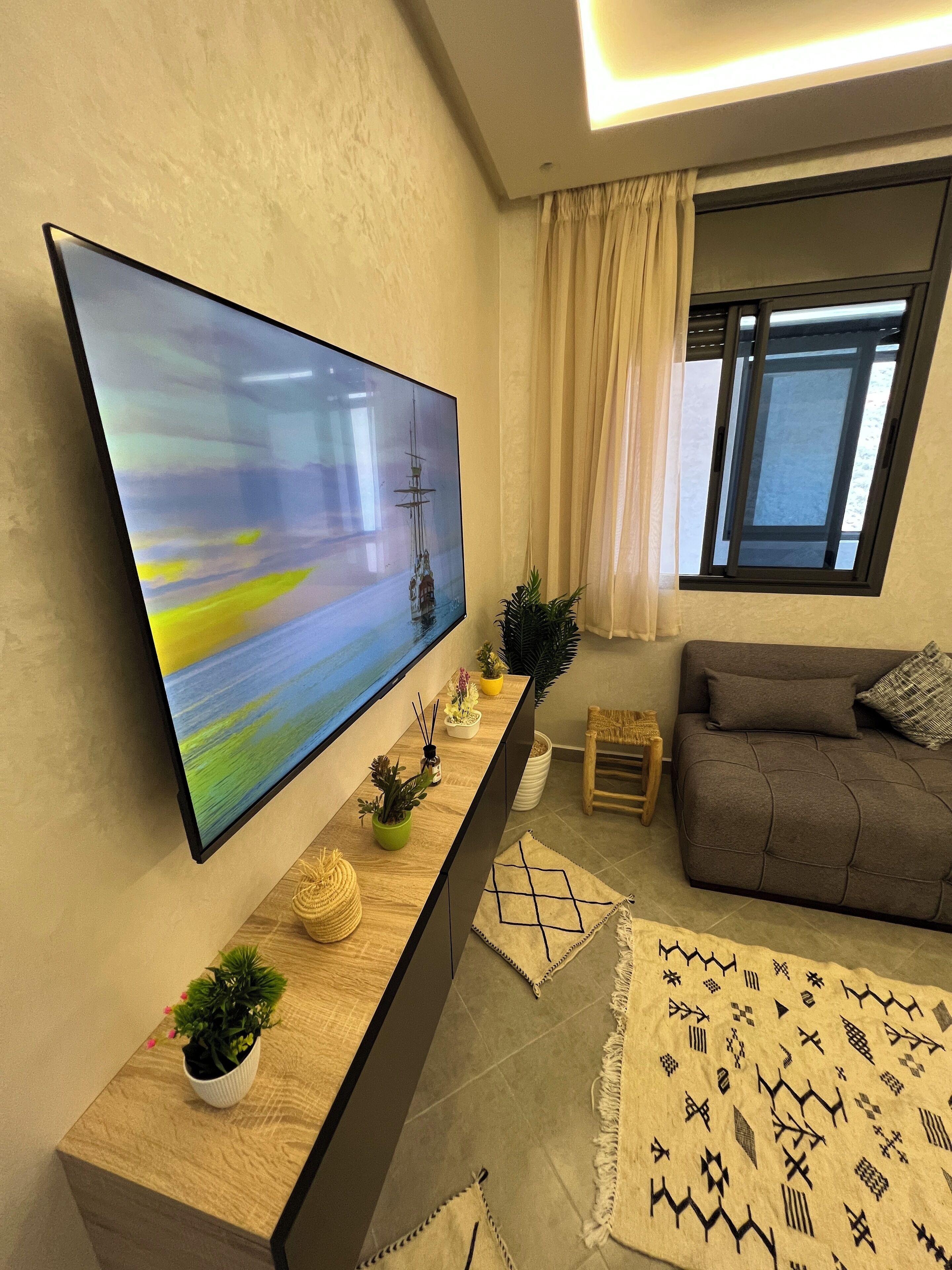 Un salon moderne doté d’un grand téléviseur à écran plat fixé au mur, d’un canapé, d’une table basse et d’une fenêtre avec des rideaux.