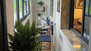 A narrow hallway with large windows, a potted plant, a small table with chairs, and a shelf with decorative items.