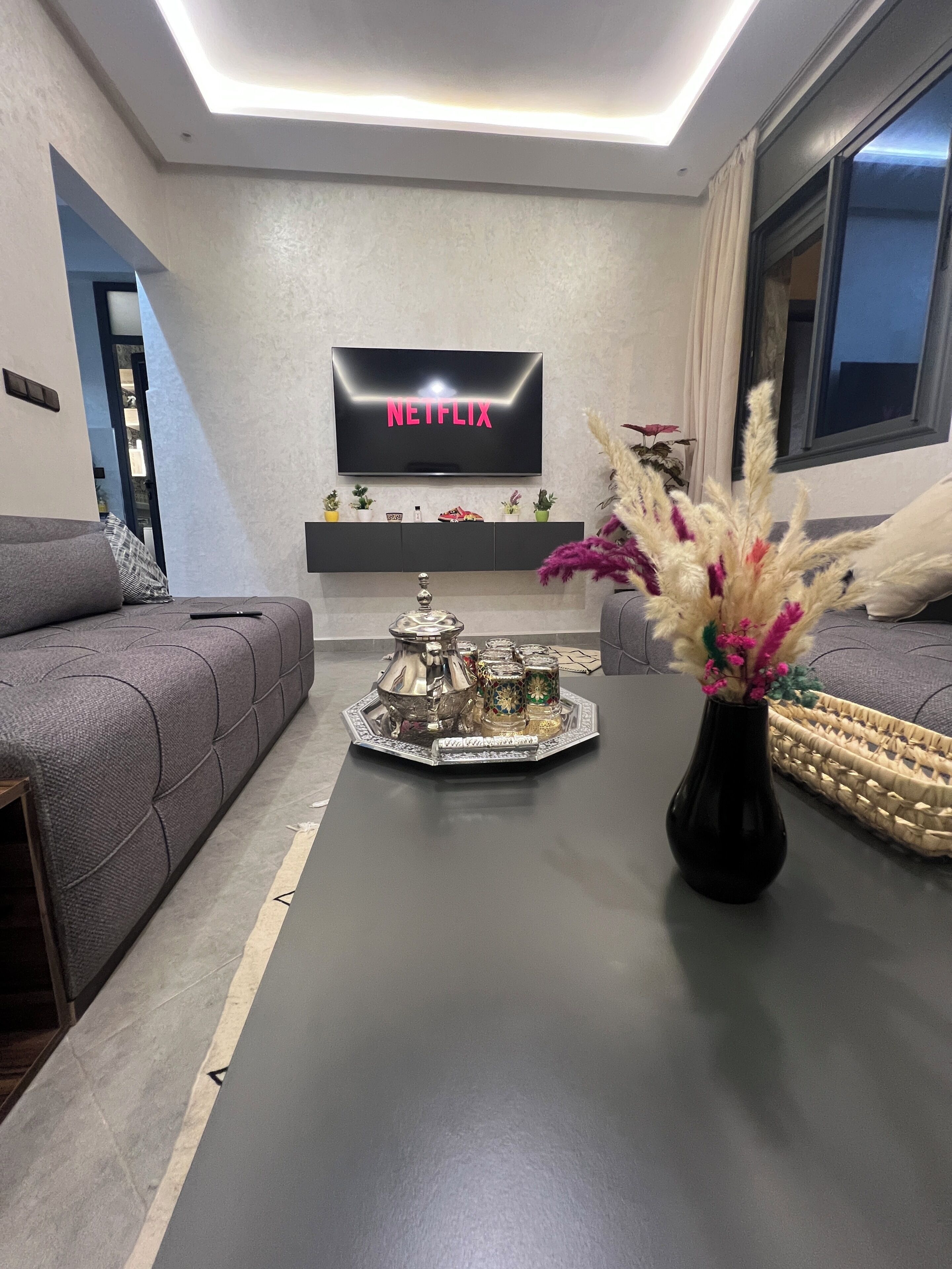 Un salon moderne avec un téléviseur à écran plat affichant le logo « NETFLIX », un canapé, une table basse agrémentée d’objets décoratifs et un vase rempli de fleurs.