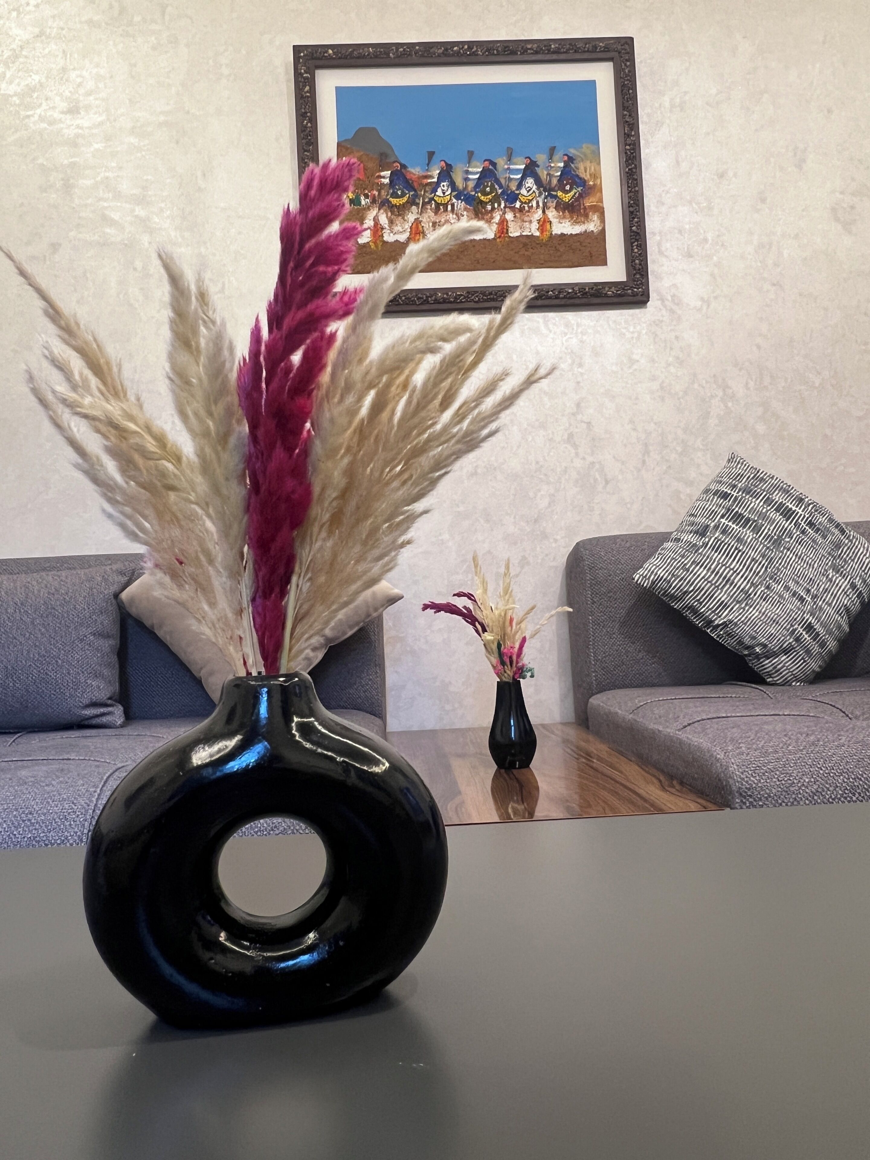 A room with a black vase on a table, a grey sofa, and a framed picture on the wall.