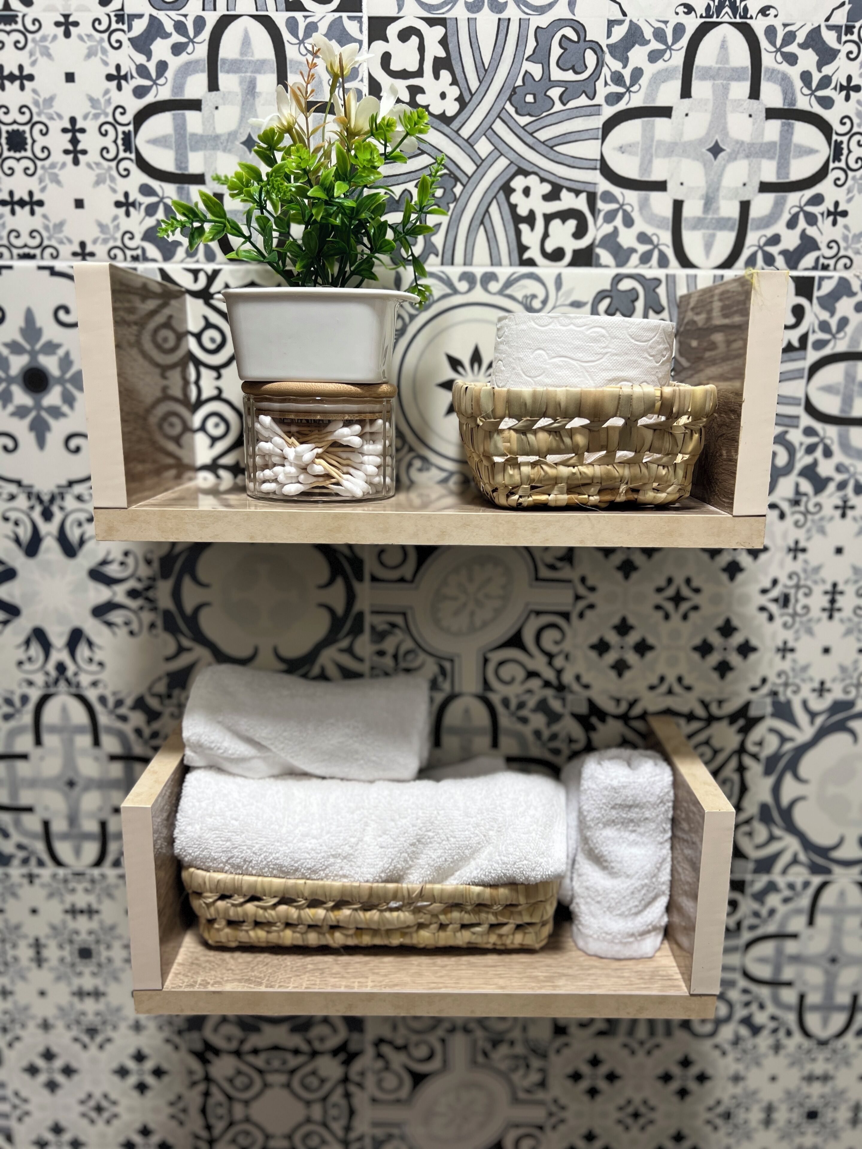 Une étagère avec un bocal en verre, un panier tressé et des serviettes pliées, le tout adossé à un mur à motifs.