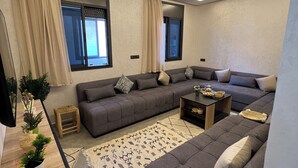 A modern living room with a grey sectional sofa, a coffee table, and a patterned rug. There are two windows with curtains, a ceiling fan, and a flat-screen TV mounted on the wall.