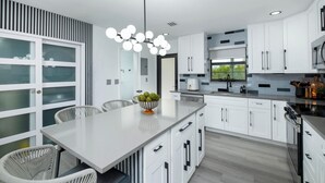 A modern kitchen with white cabinetry, a central island, and a dining area with wicker chairs. - Tropical 2BR/2BA Getaway in Gorgeous Key Largo, Pool, Kayak, Lagoon, Marina, (Key Largo)
