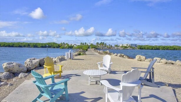 A beachside seating area with Adirondack chairs and a concrete table, overlooking a calm body of water and a distant shoreline with buildings. - Tropical 2BR/2BA Getaway in Gorgeous Key Largo, Pool, Kayak, Lagoon, Marina, (Key Largo)